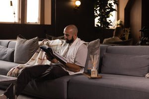 Man on a sofa reading next to a reed diffuser and blanket