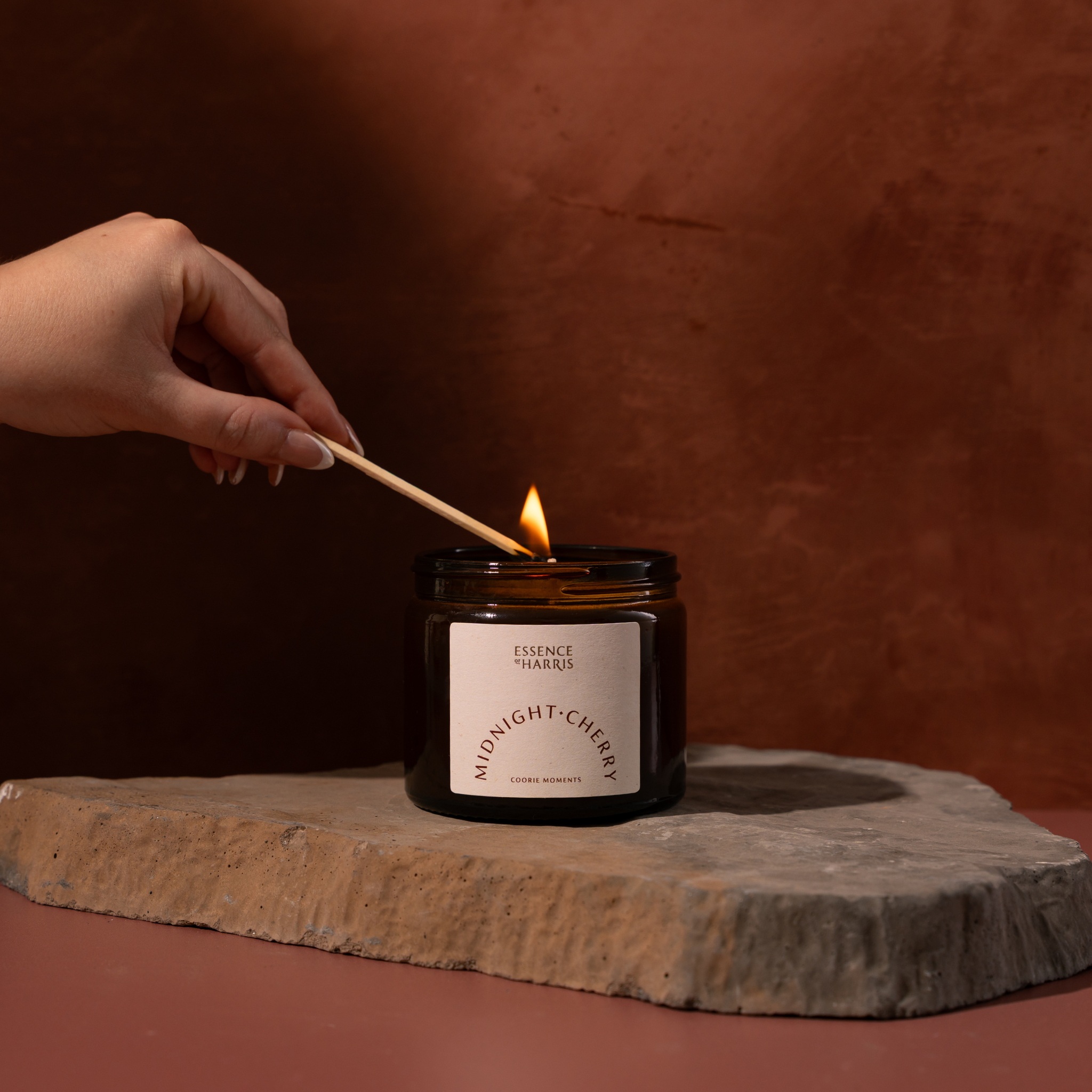 A hand holds a lit match over an amber glass jar candle labeled 'ESSENCE HARRIS' and 'MIDNIGHT CHERRY' on a rustic stone slab against a warm terracotta background.