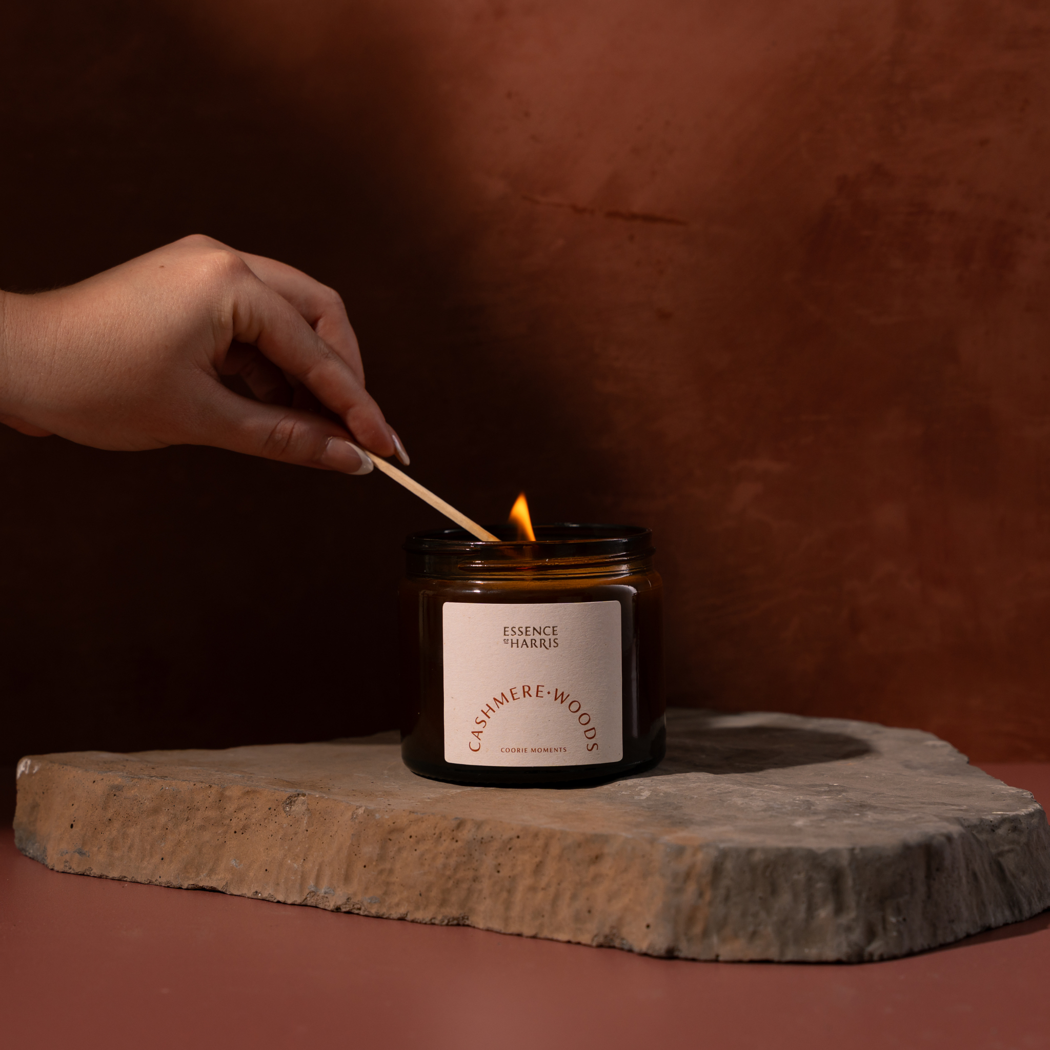 A hand holds a wooden match above a lit amber glass candle jar with a cream label reading Essence of Harris Cashmere Woods. The candle sits on a stone slab against a warm brown textured background.