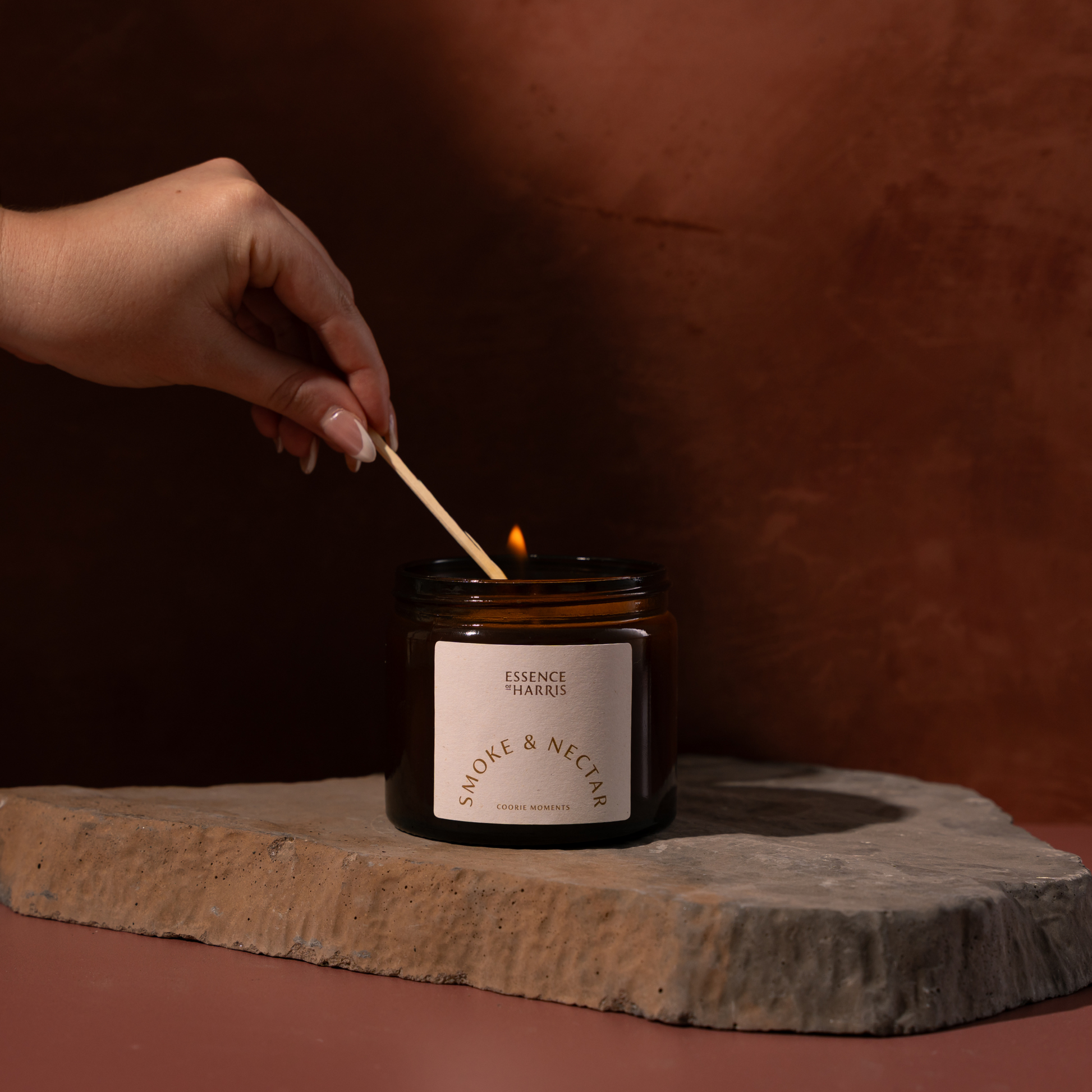 A hand holds a wooden match above a lit amber glass candle jar with a cream label reading Essence of Harris Smoke & Nectar. The candle sits on a stone slab against a warm brown textured background.