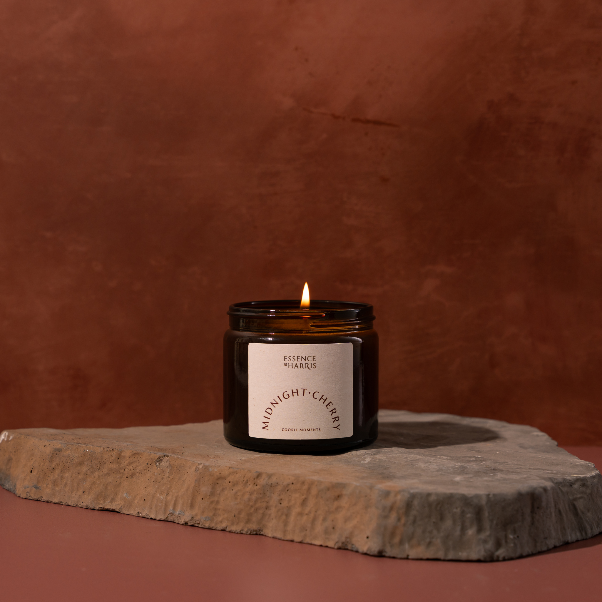 A lit amber glass jar candle with a white label reading Essence of Harris Midnight Cherry sits on a rough stone slab against a warm terracotta background.