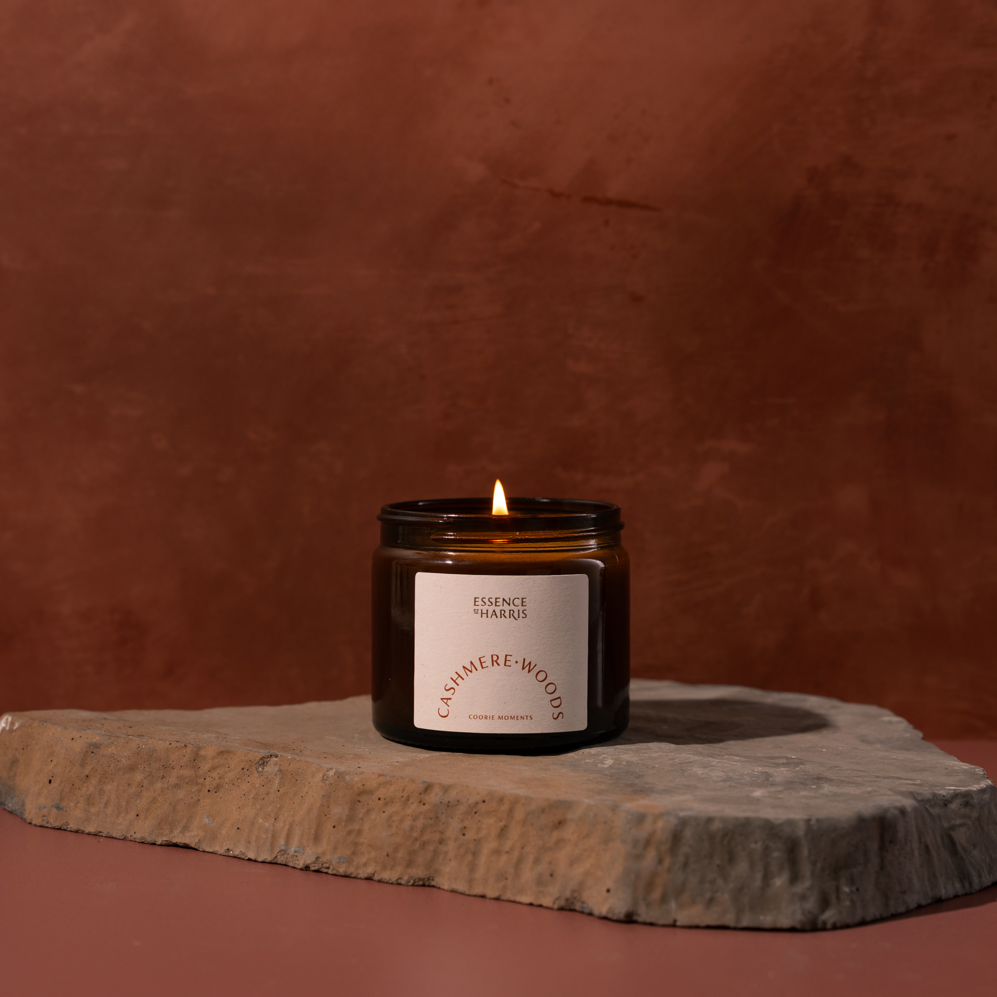 A lit amber glass jar candle with a cream label reading Essence of Harris Cashmere Woods sits on a stone slab against a warm terracotta background.