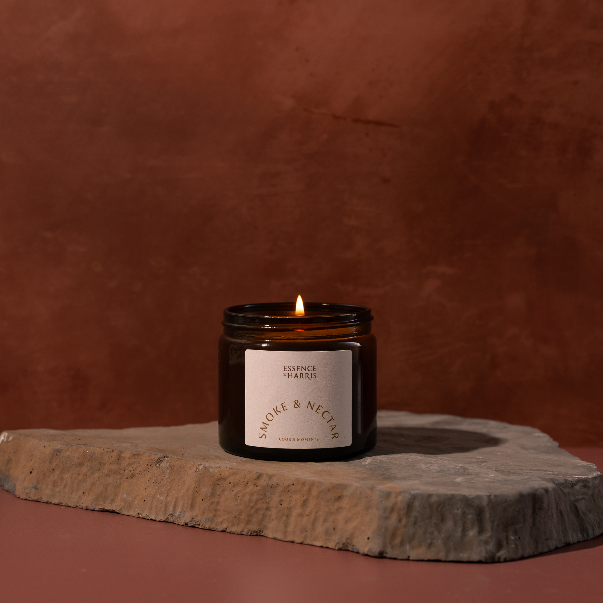 A lit amber glass jar candle with a cream label reading Essence of Harris Smoke & Nectar sits on a stone slab against a warm terracotta background.