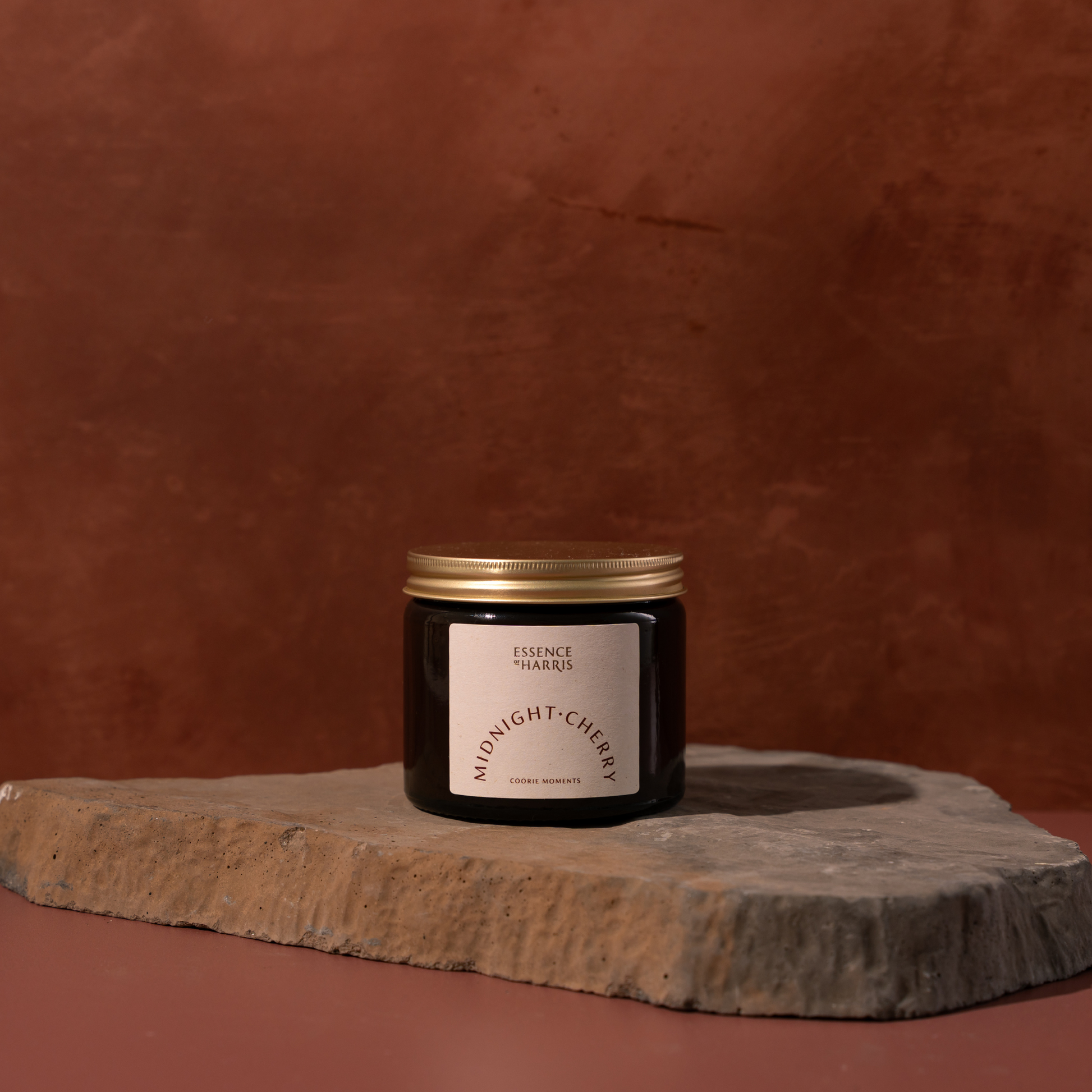 A dark glass jar candle with a gold lid and a white label reading 'ESSENCE OF HARRIS' and 'MIDNIGHT CHERRY' sits on a rough stone slab against a warm terracotta background.