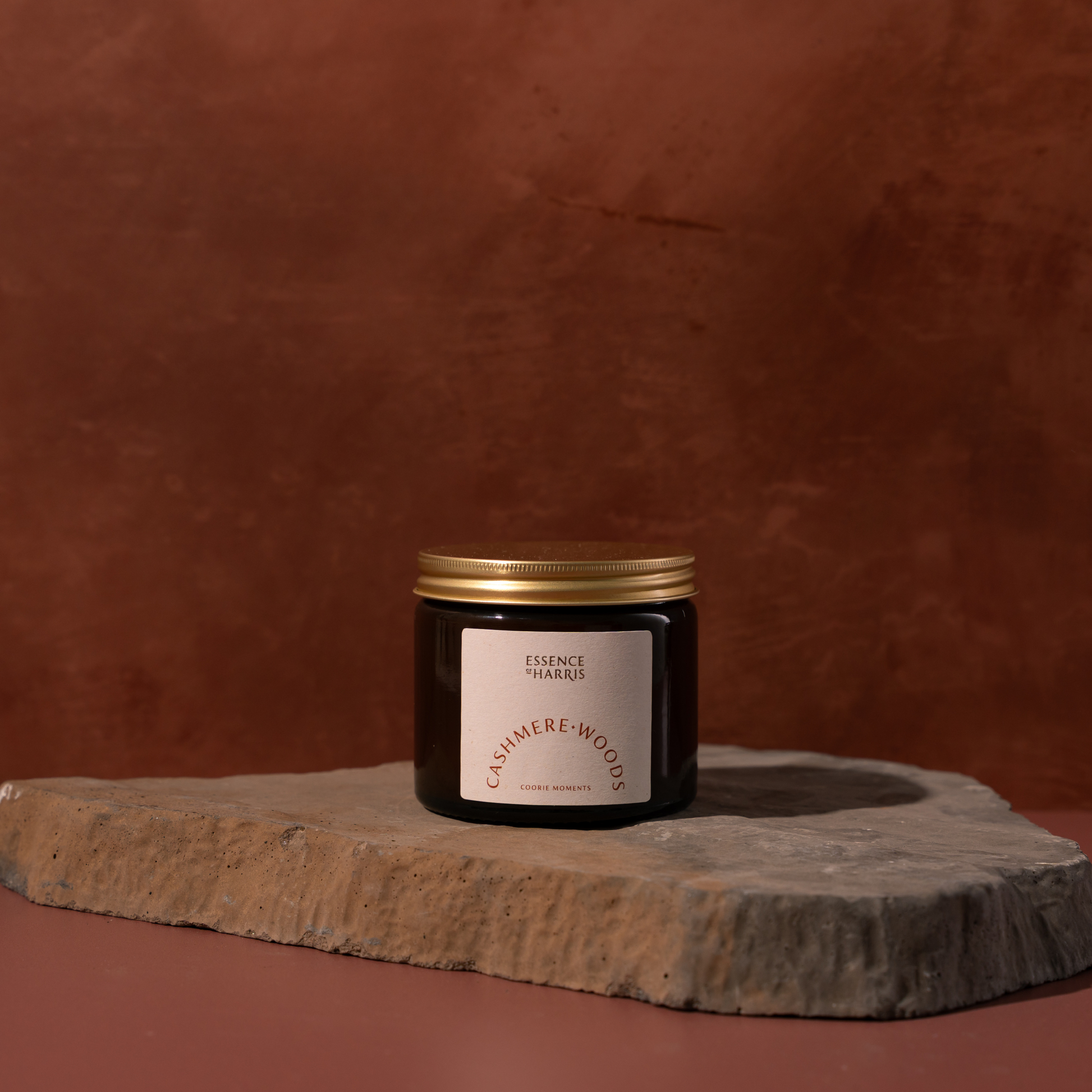 A dark amber glass jar candle with a gold metal lid and a white label reading Essence of Harris Cashmere Woods, placed on a rough stone slab against a warm terracotta background.