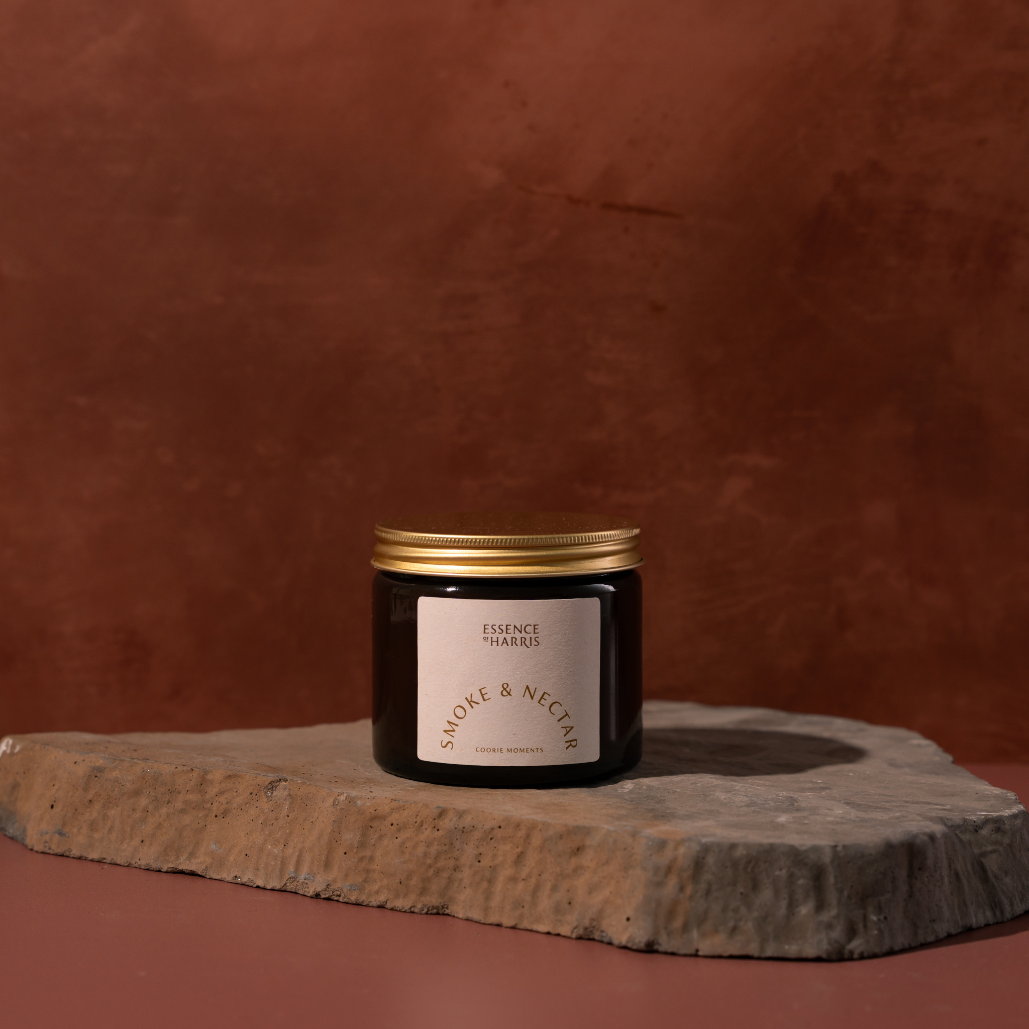 A dark amber glass jar candle with a gold metal lid and a cream label reading Essence of Harris Smoke & Nectar, displayed on a rough stone slab against a warm terracotta background.
