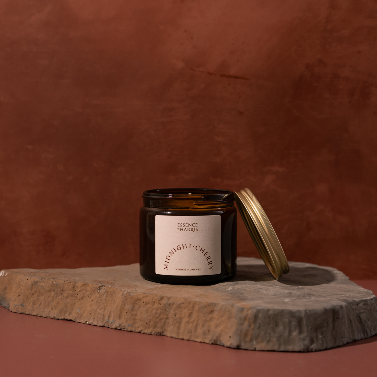 An amber glass candle jar with a cream label reading Essence of Harris Midnight Cherry sits on a rough stone slab against a warm terracotta background, with its gold lid leaning beside it.