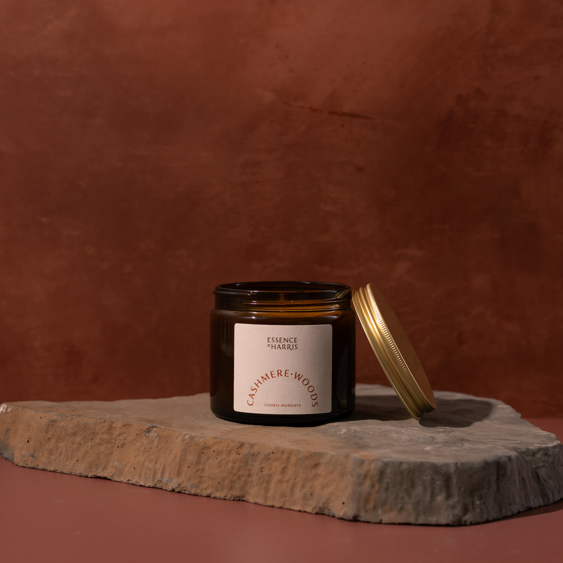 An amber glass candle jar with a cream label reading Essence of Harris Cashmere Woods sits on a rough stone slab against a warm terracotta background, with its gold lid leaning beside it.