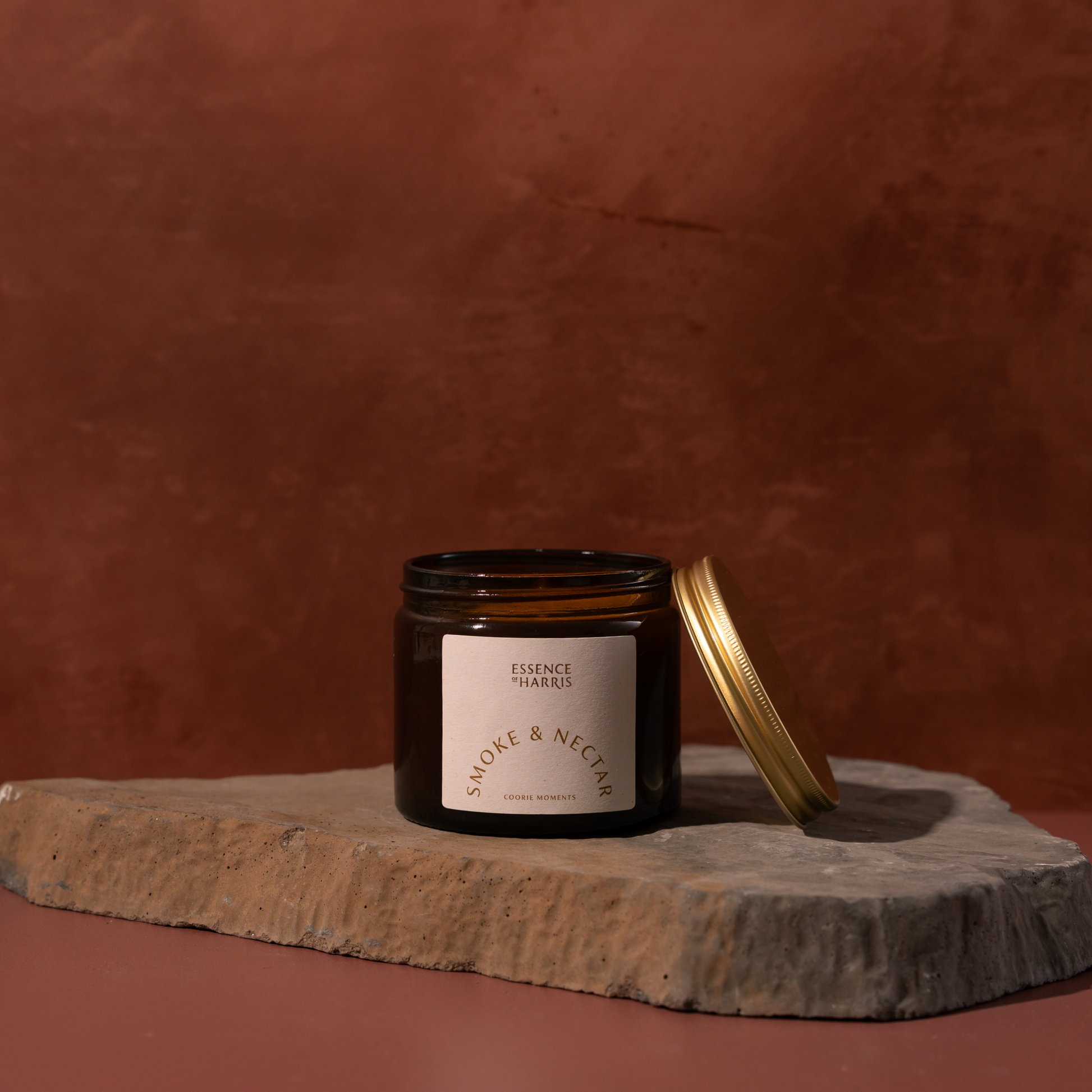 An amber glass candle jar with a cream label reading Essence of Harris Smoke & Nectar sits on a rough stone slab against a warm terracotta background, with its gold lid leaning beside it.