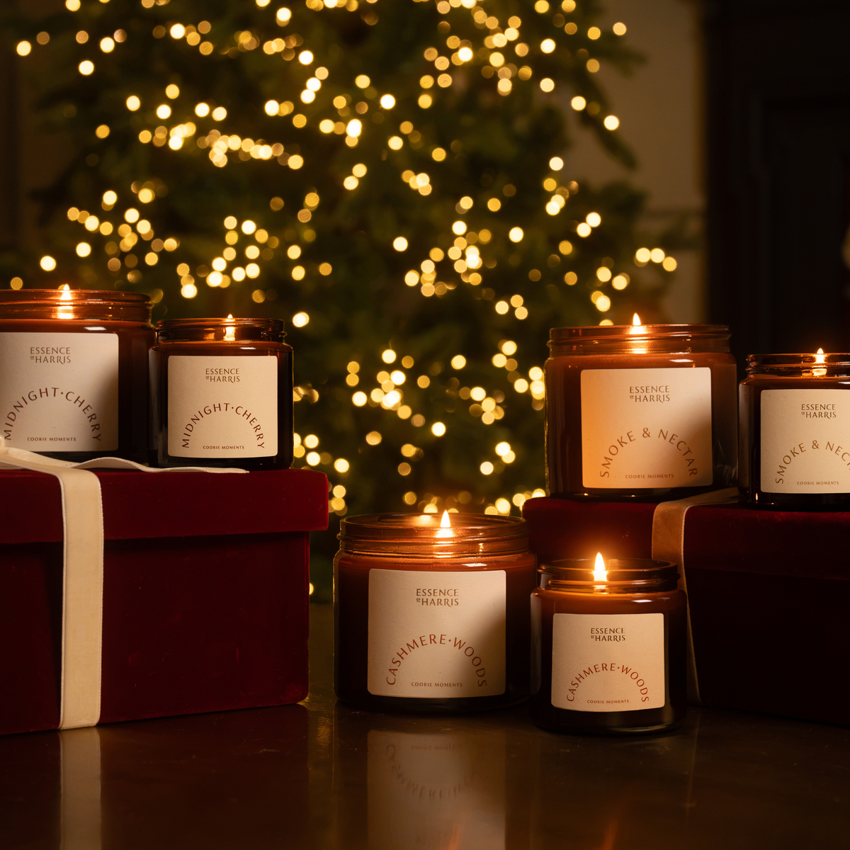 A collection of lit glass jar candles from Essence of Harris arranged on a wooden table in front of a blurred Christmas tree with warm white Christmas lights. The candles come in dark amber glass jars with white labels showing scents like Midnight Cherry, Cashmere Woods, and Smoke & Nectar.