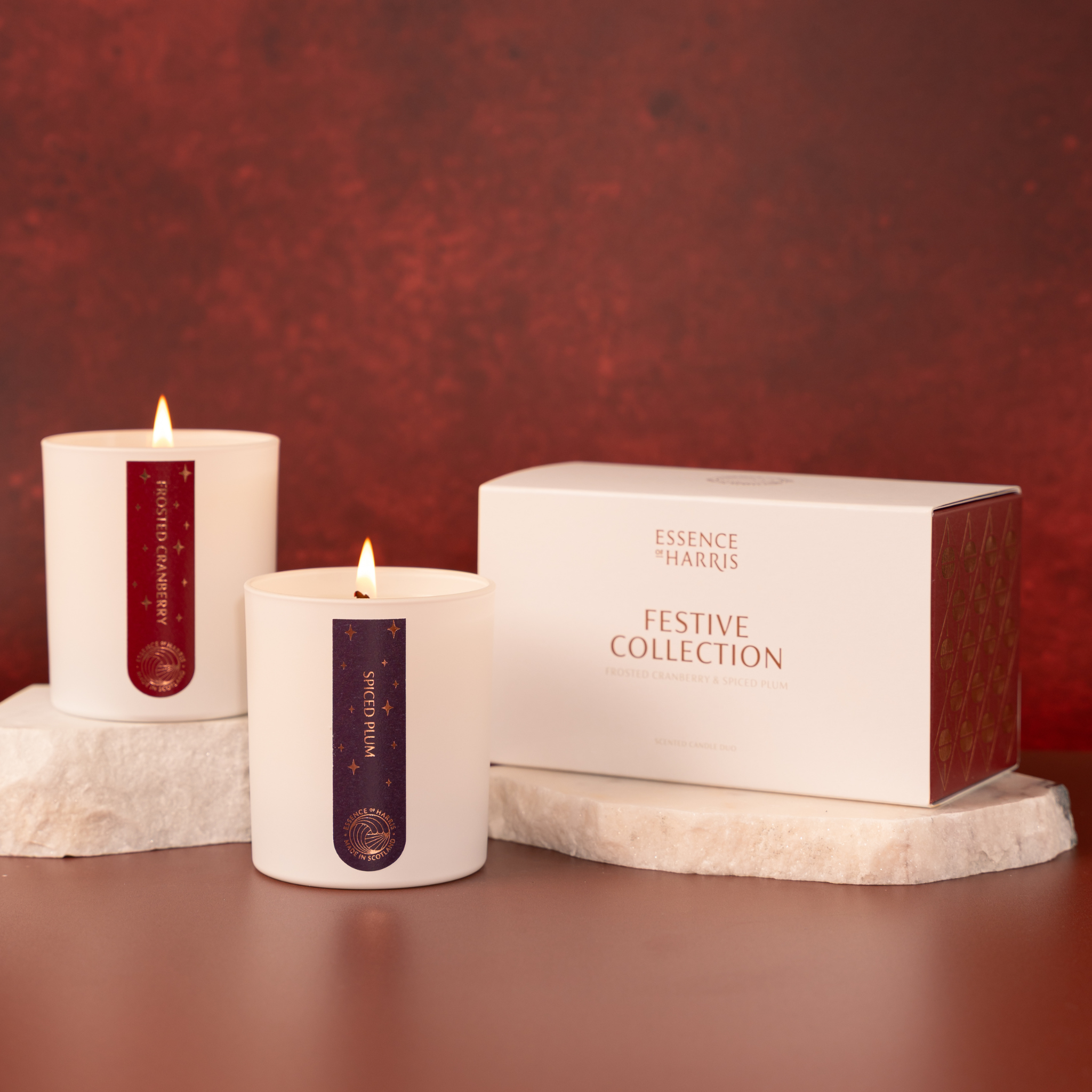 Essence of Harris Frosted Cranberry and Spiced Plum candles with lit flames and a white gift box labeled 'Essence of Harris Festive Collection' on a warm brown surface against a textured dark red background.