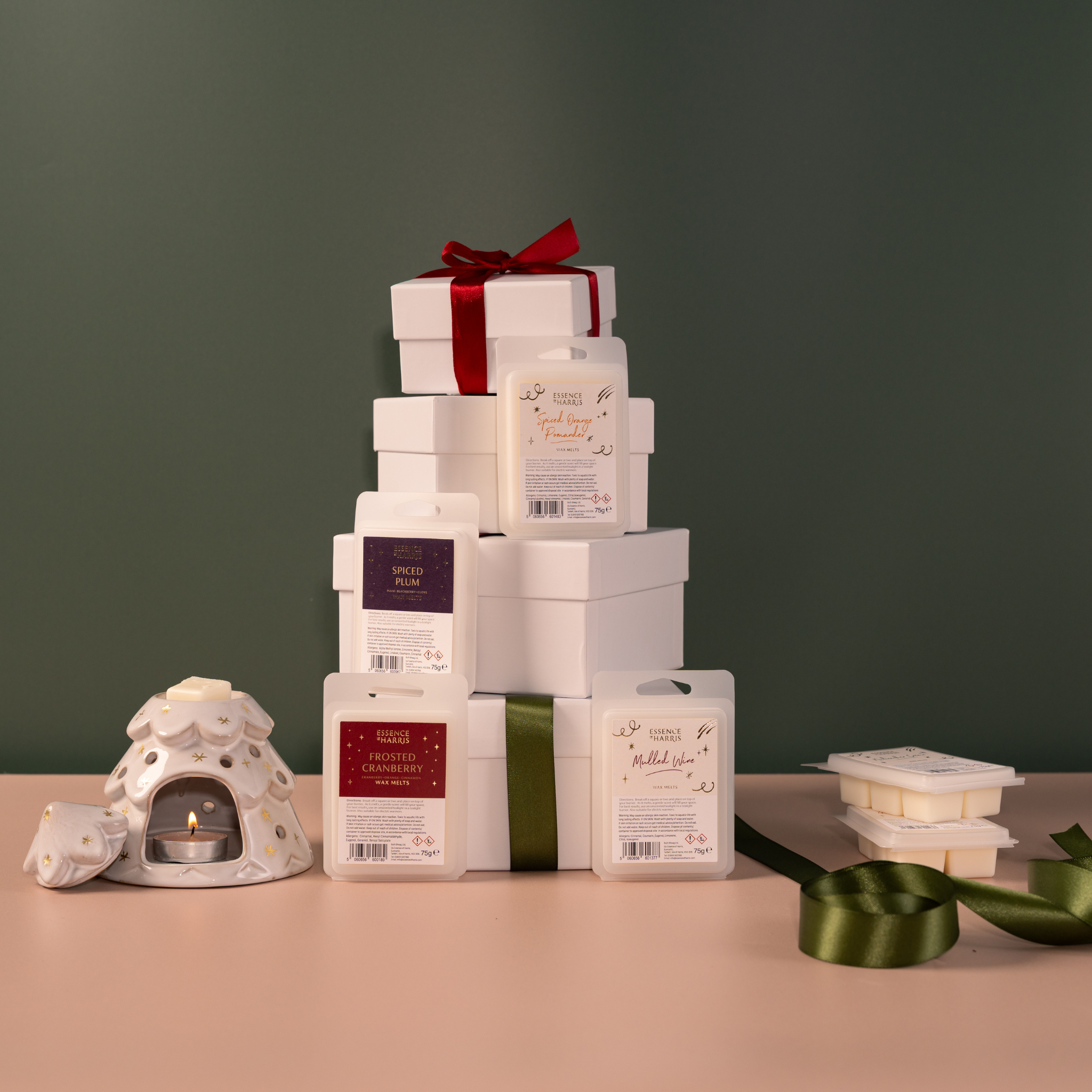 A stack of gift boxes and wax melts bars arranged on a tabletop against a dark green wall, with a small white ceramic tealight candle holder, green ribbon, and additional wax melts in the foreground.