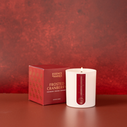 A white glass candle with a lit flame sits next to its red packaging box. The candle features a vertical red label with gold text reading 'Essence of Harris' and 'Frosted Cranberry'.