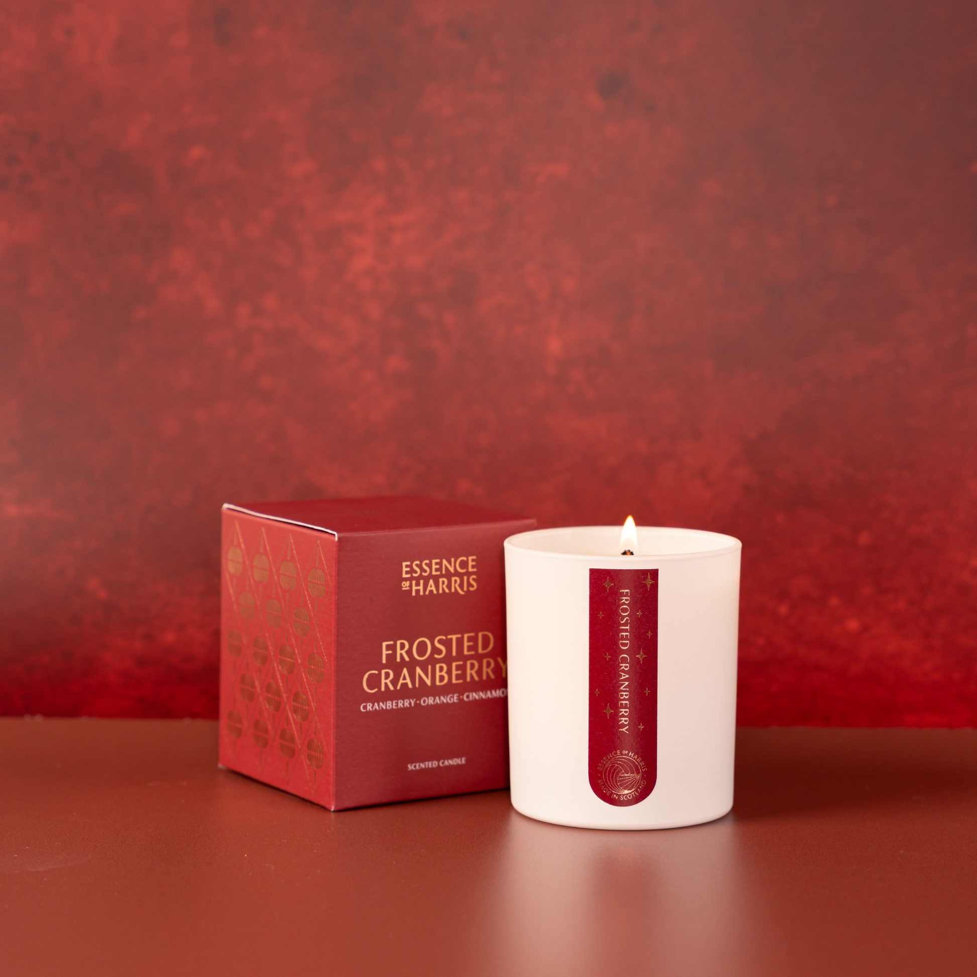 A white glass candle with a lit flame sits next to its red packaging box. The candle features a vertical red label with gold text reading 'Essence of Harris' and 'Frosted Cranberry'.
