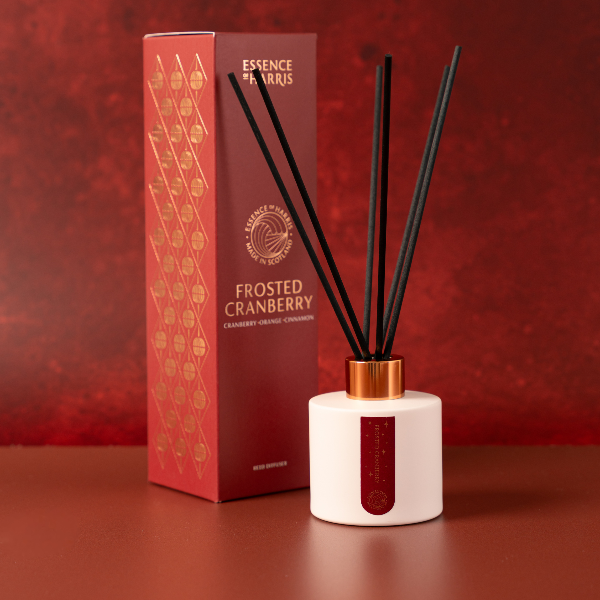 Essence of Farris Frosted Cranberry reed diffuser with a white cylindrical bottle, black reeds, and a metallic gold cap, shown next to its red and gold packaging box on a matching red surface.