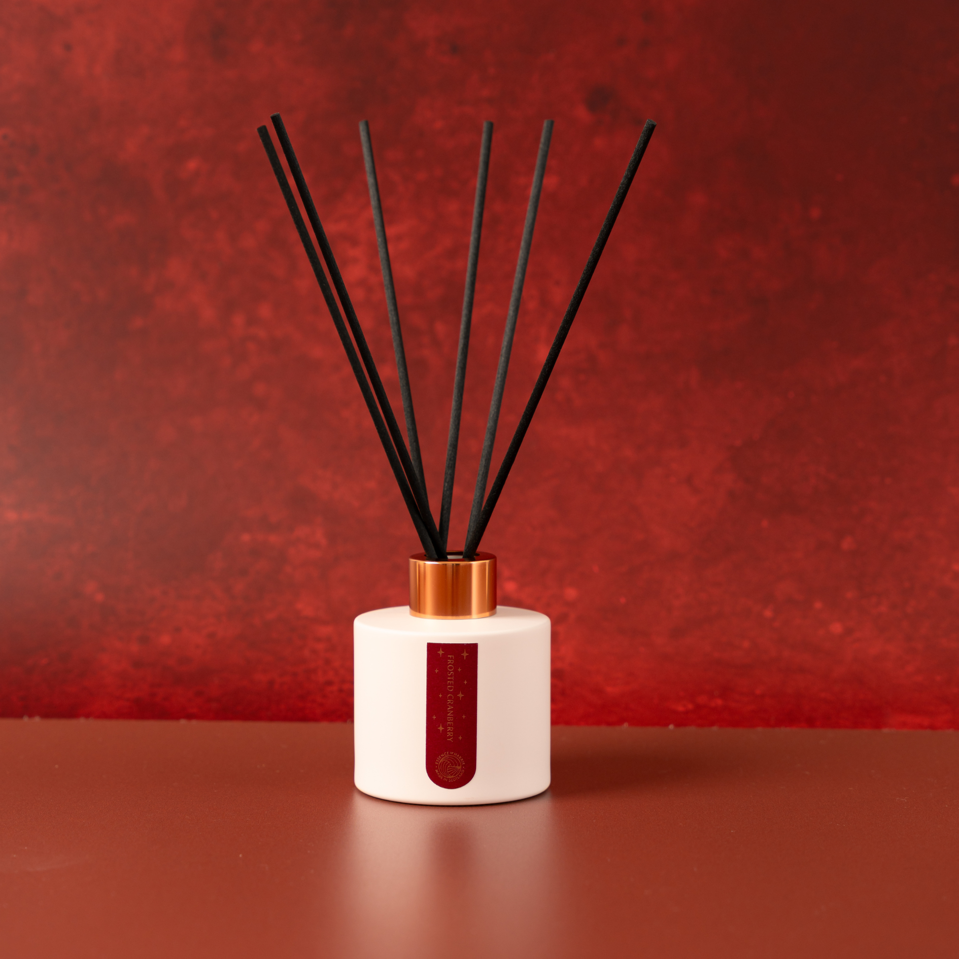Essence of Harris Frosted Cranberry reed diffuser with a white cylindrical bottle, a metallic copper collar, and six black reed sticks, set against a textured red background.