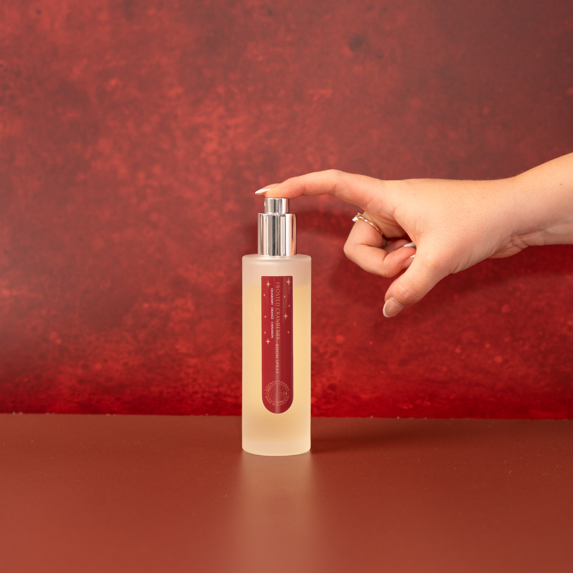 Frosted Cranberry Room Spray in frosted glass with silver spray cap photographed on a red festive background. With manicured hand pressing down on spray cap.