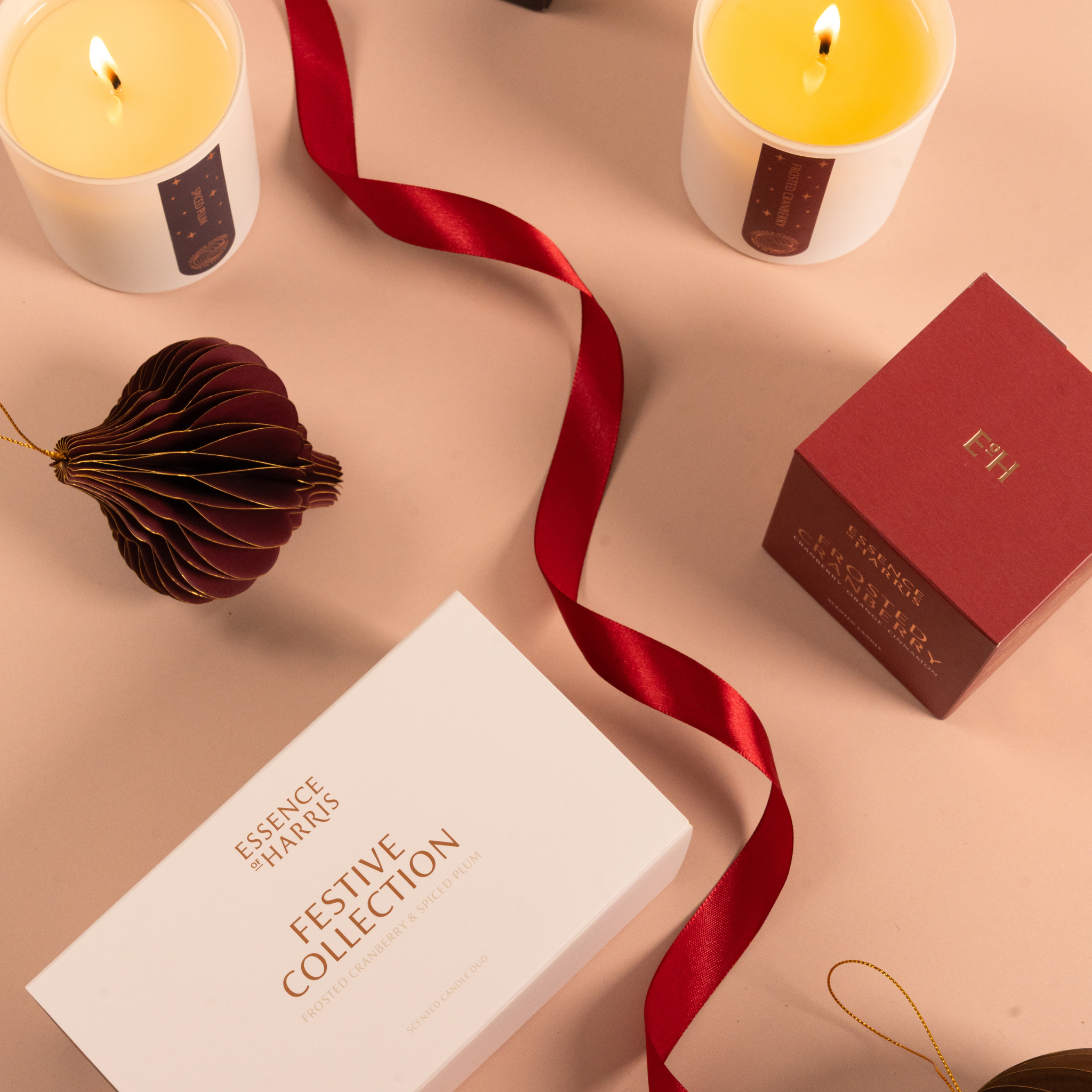 A styled flat-lay of the Essence Harris Festive Collection, featuring a white candle gift box labeled 'Festive Collection' and a red box labeled 'Frosted Cranberry' on a warm beige background, accented by a red ribbon, lit candles, and a decorative paper ornament.