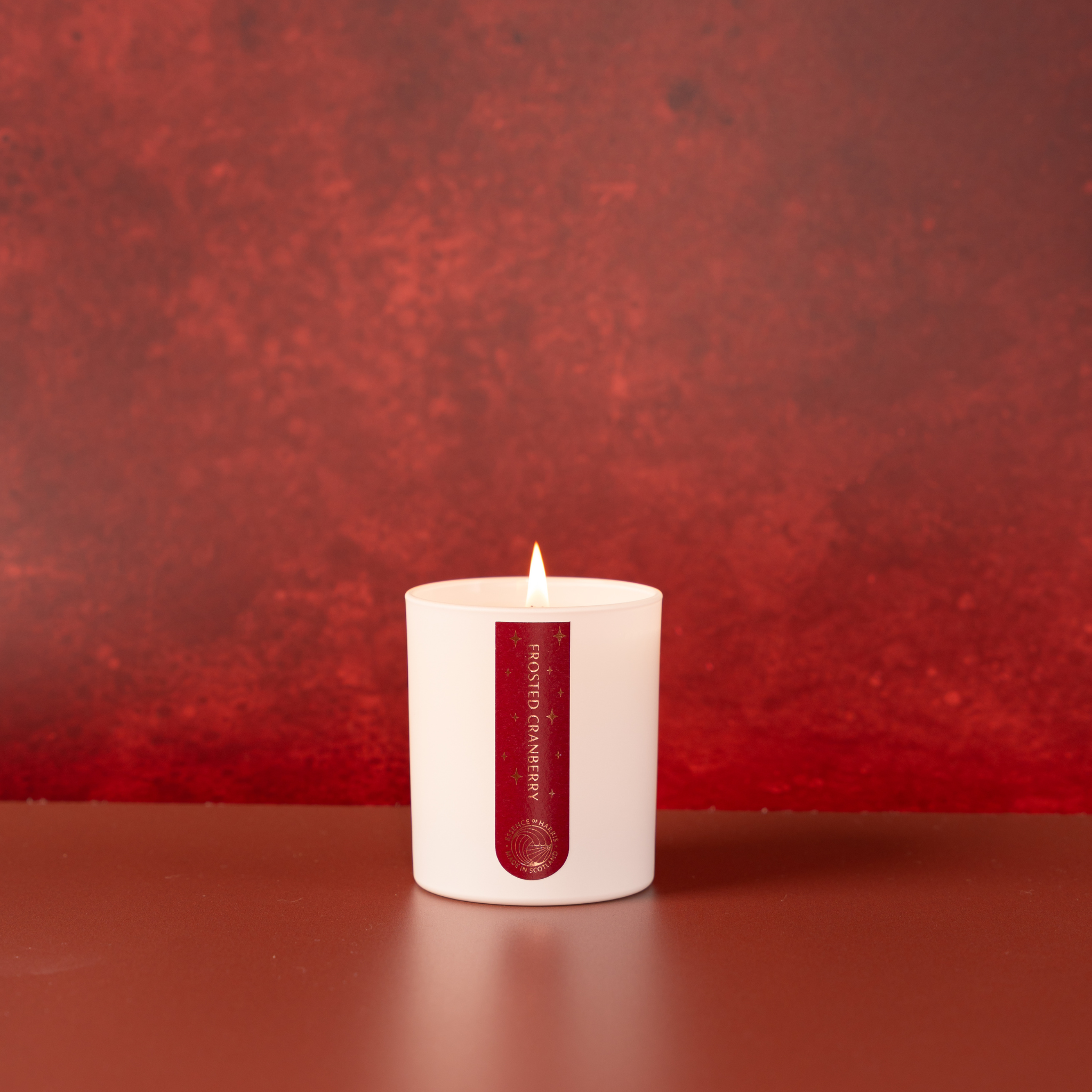 A white jar candle with a lit wick sits centered on a dark red surface against a textured red background. The front of the jar has a vertical dark red label with gold foiled text reading 'Frosted Cranberry' and a small circular Essence of Harris logo near the bottom.