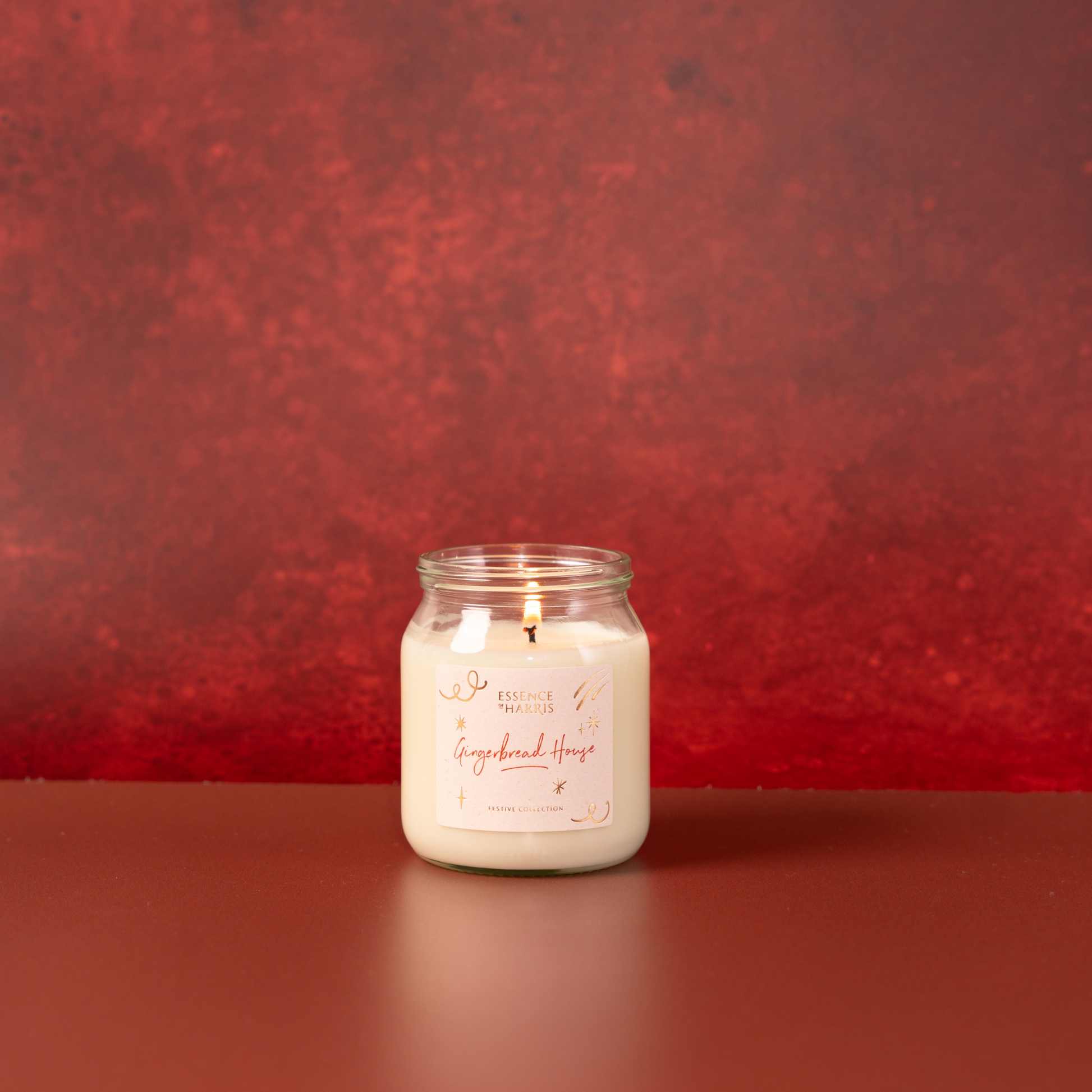 A lit white soy candle in a clear glass jar with a white label reading 'Gingerbread House', set against a textured red festive background.
