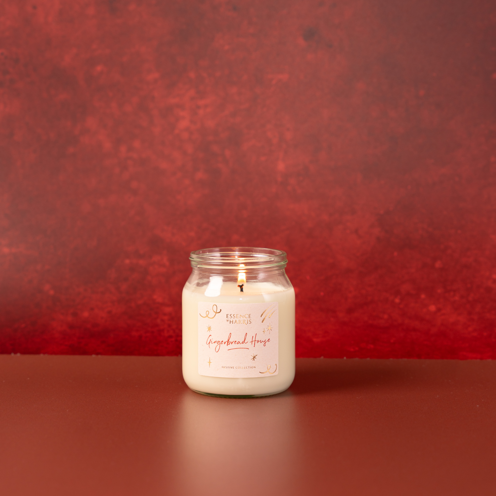 A lit white soy candle in a clear glass jar with a white label reading 'Gingerbread House', set against a textured red festive background.