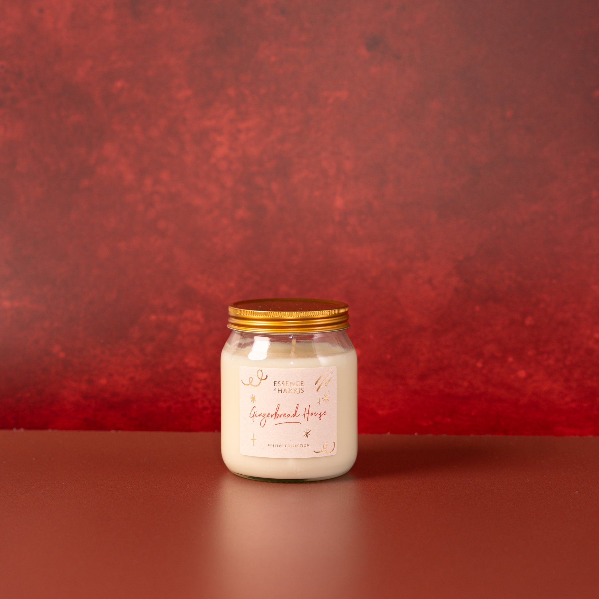 A glass jar candle with a gold lid and cream-colored wax, featuring a label that reads 'Gingerbread House', set against a textured red background.