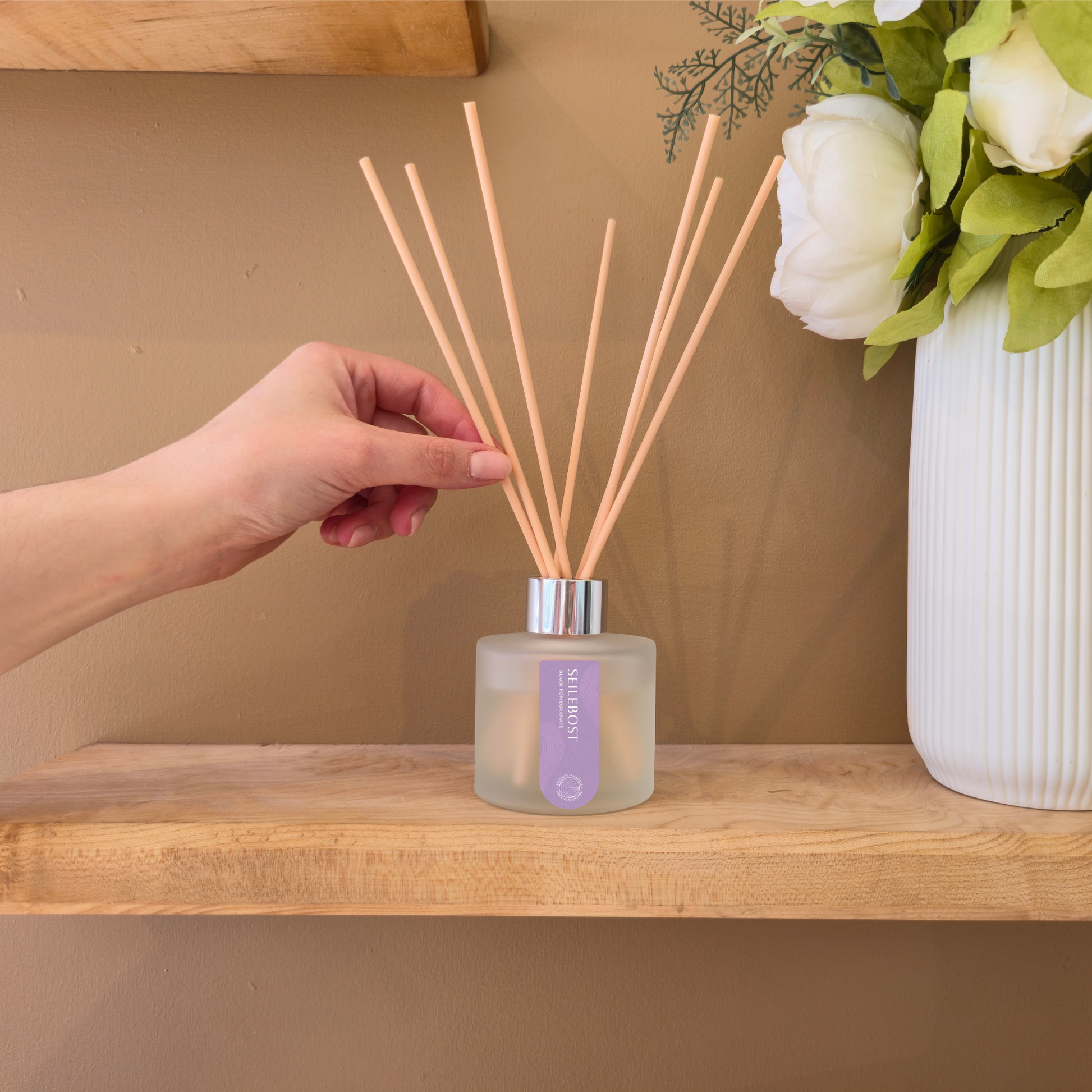 Hand holding a essence of harris seilebost diffuser with reeds on a wooden shelf with a vase of flowers in the background