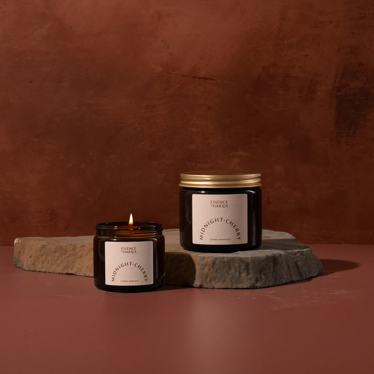 Two dark amber glass candles with white labels are displayed on a stone slab against a warm rust-brown background. One candle is lit and sits in front of the other, which has a gold metal lid.