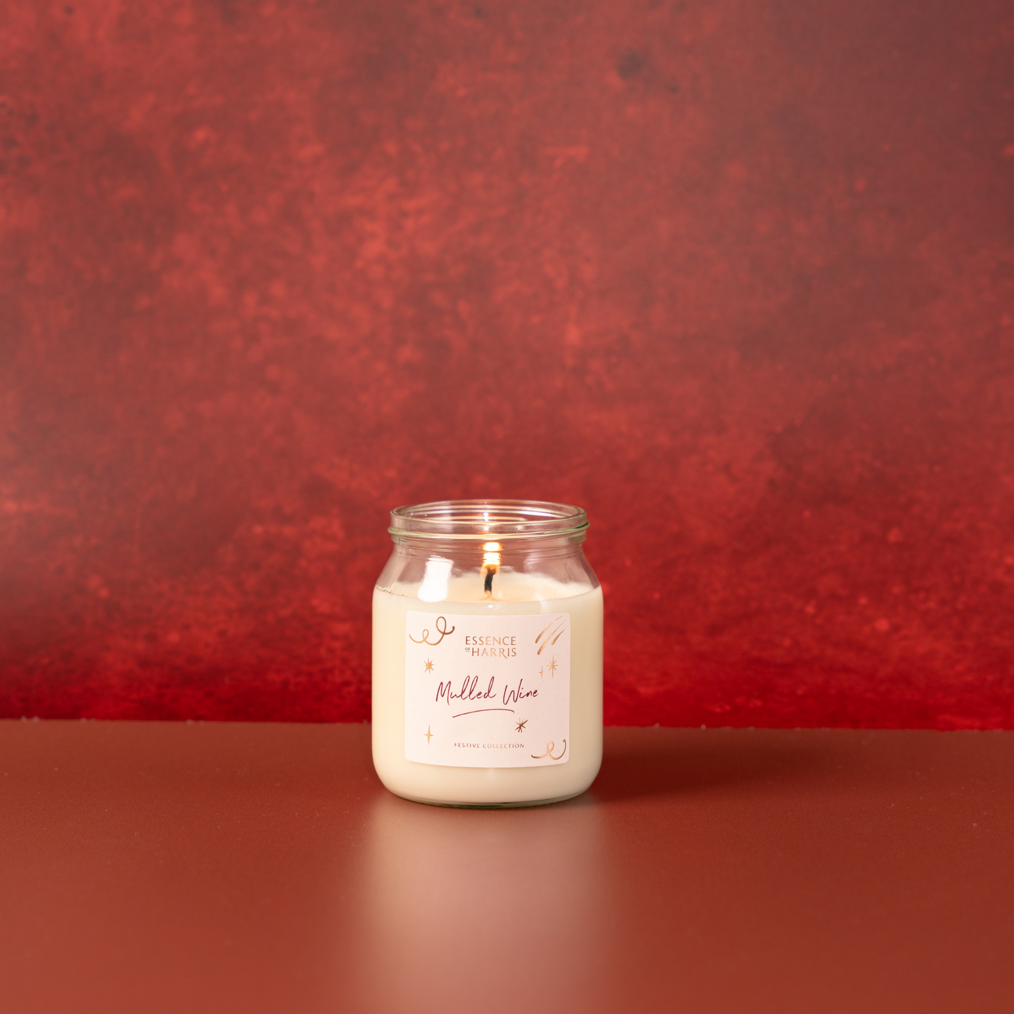 A lit candle in a clear glass jar with white wax sits against a textured red background. The front label reads 'Mulled Wine'.