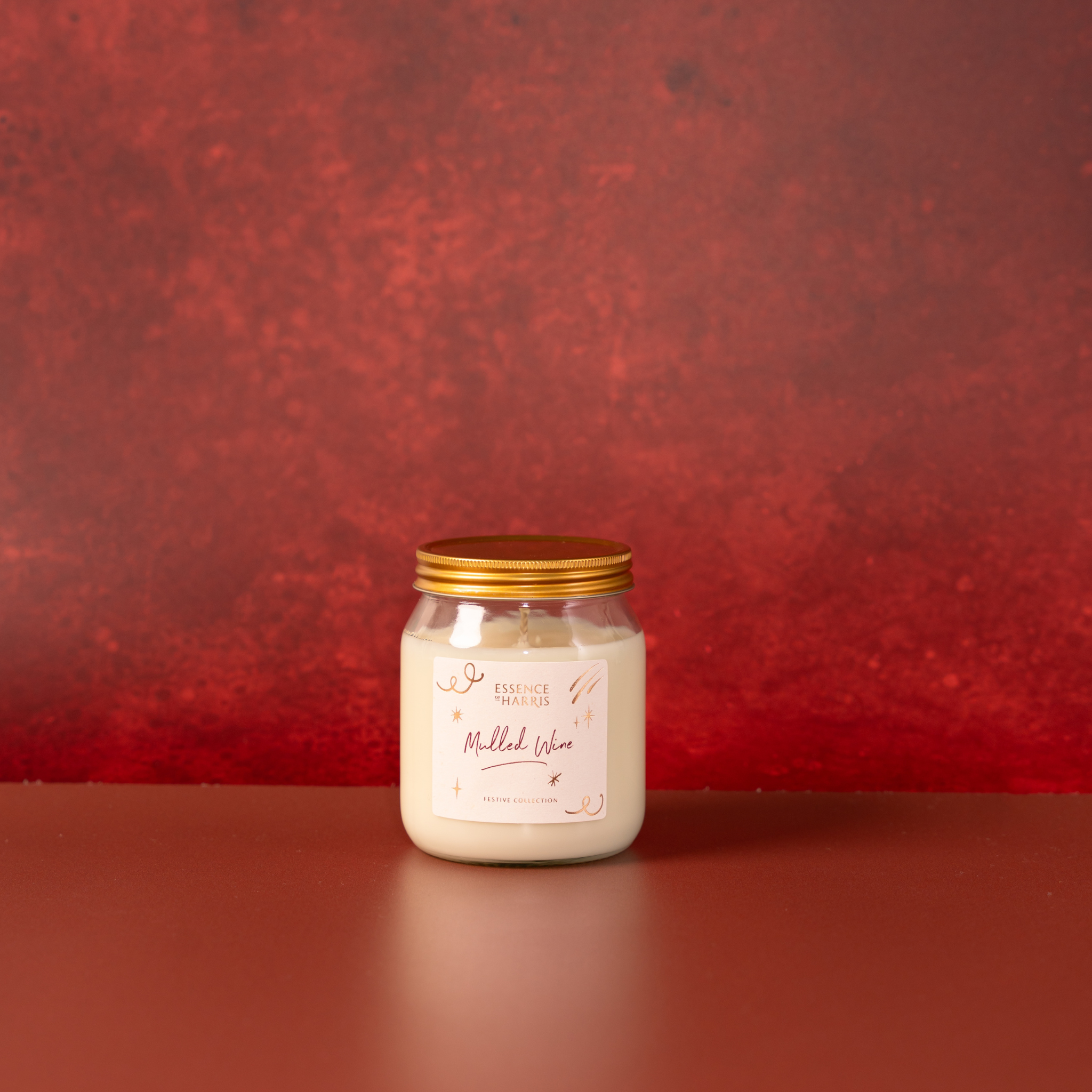 A glass jar candle with a gold metal lid and a cream-colored label reading 'Mulled Wine' on a matching brown surface against a textured red background.