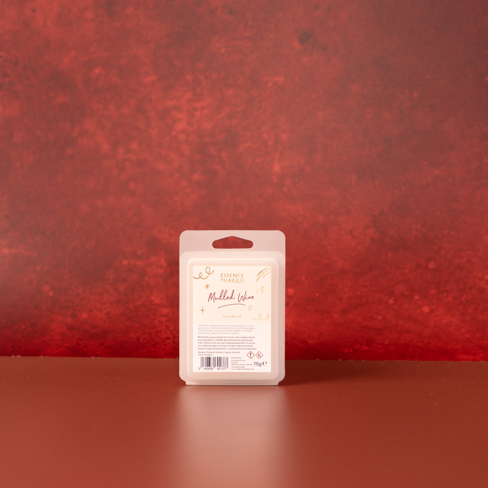 A single packaged wax melt bar stands upright on a smooth reddish-brown surface against a textured red wall. The package is a frosted translucent plastic clamshell with a cream label reading 'Mulled Wine'.