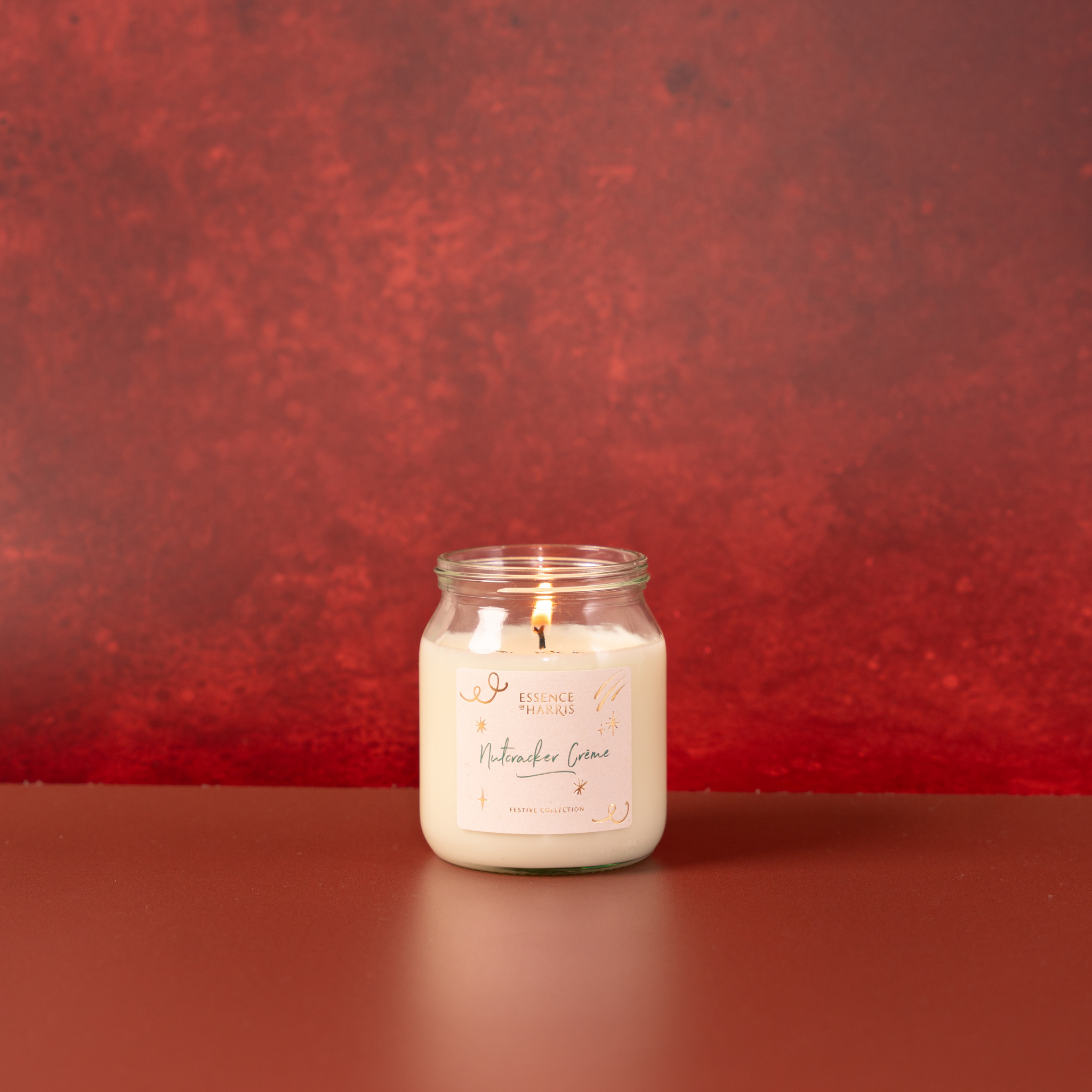 A lit scented candle in a clear glass jar sits on a dark reddish-brown surface against a textured red background. The label reads 'Nutcracker Creme'.