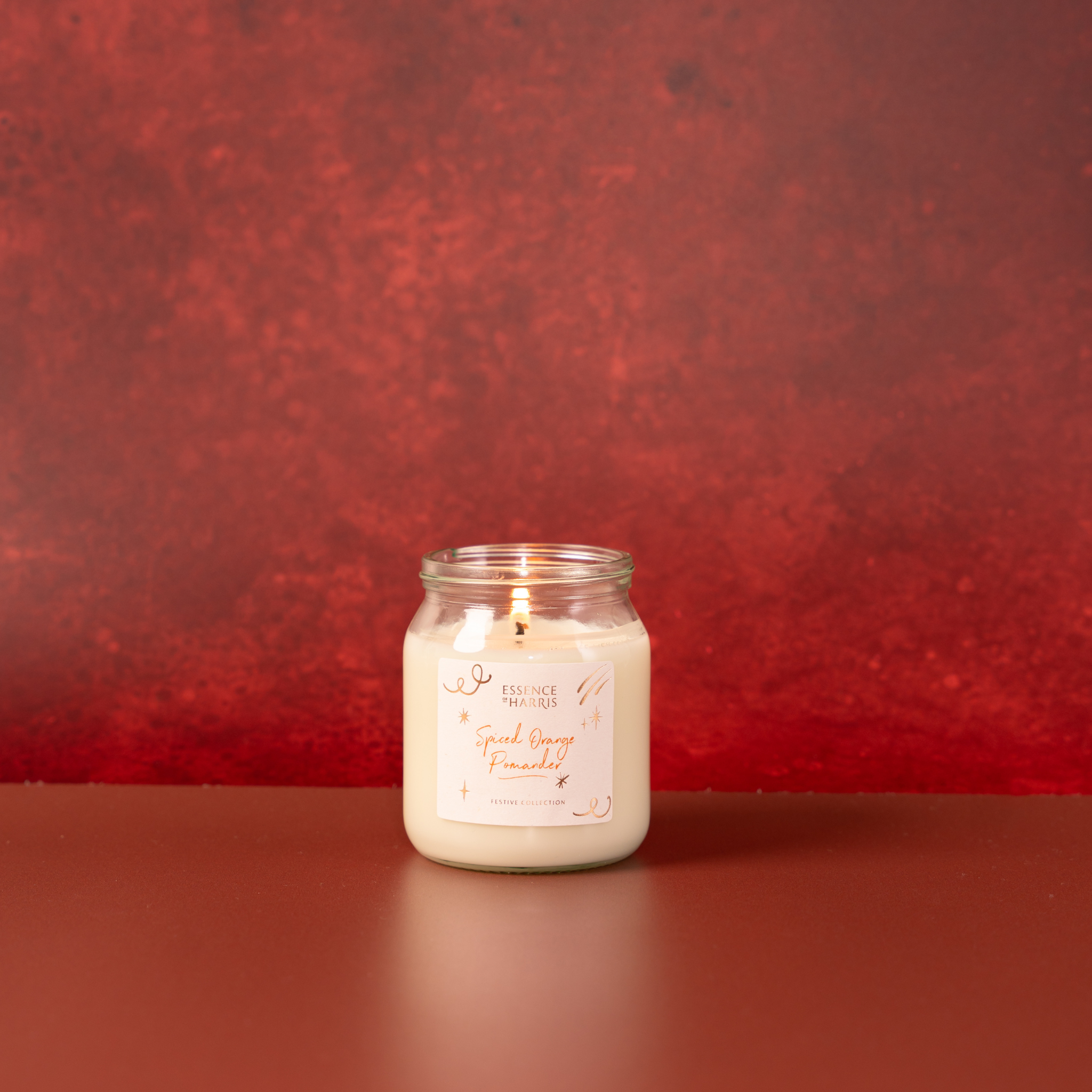 Essence of Harris Spiced Orange Pomanger lit jar candle with white wax sits on a dark red festive surface. The glass jar has a white label with elegant script text and gold foiling.