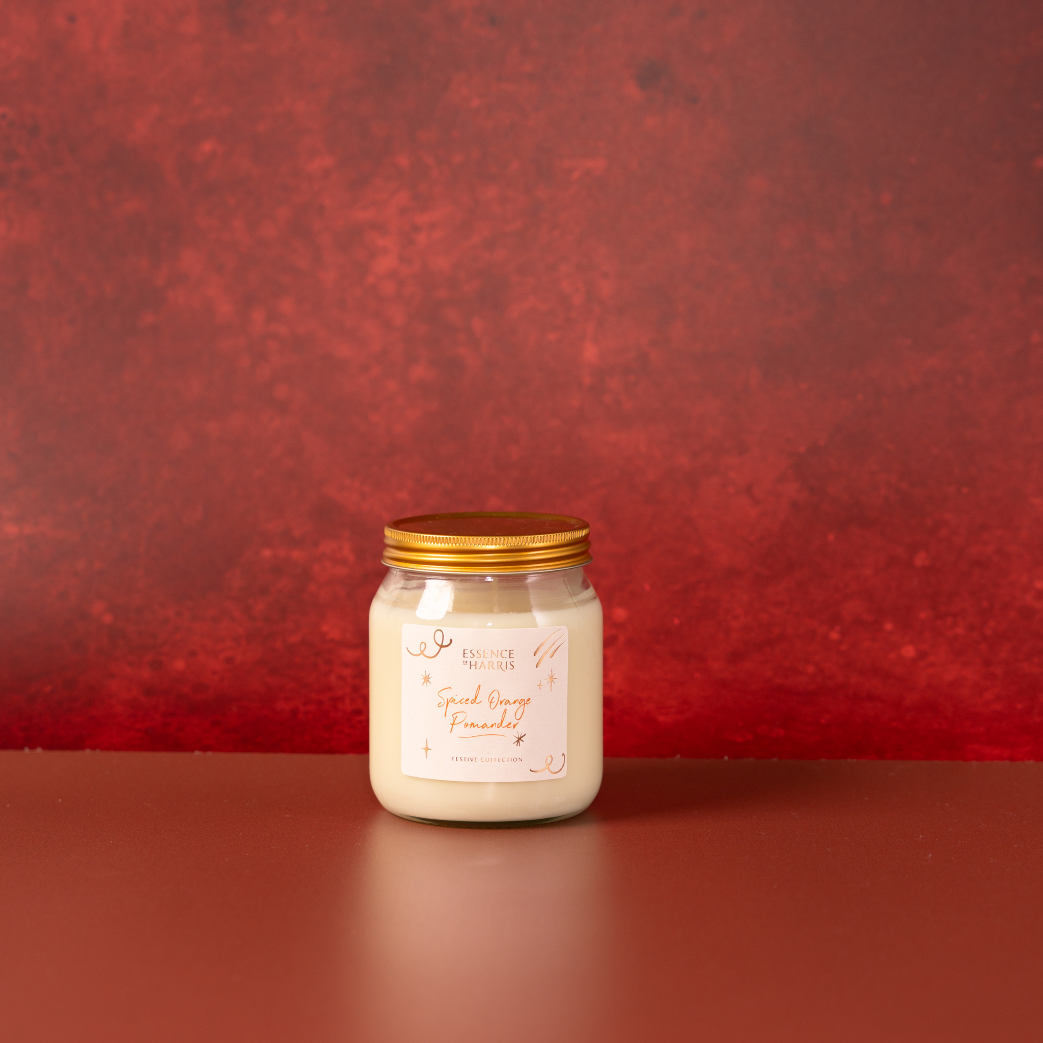 Essence of Harris Spiced Orange Pomanger jar candle with gold lid and white wax on a dark red festive surface. The glass jar has a white label with elegant script text and gold foiling.