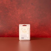 A packaged wax melt bar stands upright on a reddish festive surface against a textured red wall. The package is a beige plastic clamshell with a white front label reading 'Spiced Orange Pomander' in gold script.