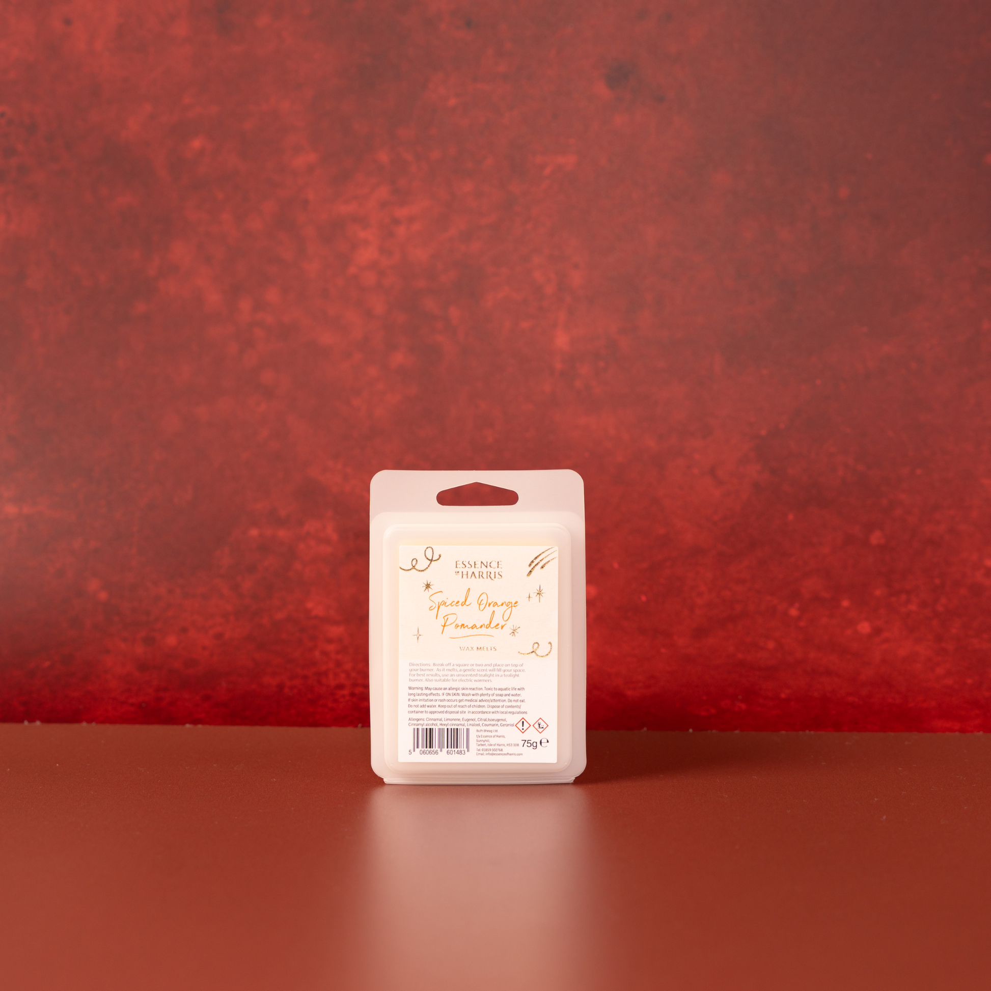A packaged wax melt bar stands upright on a reddish festive surface against a textured red wall. The package is a beige plastic clamshell with a white front label reading 'Spiced Orange Pomander' in gold script.