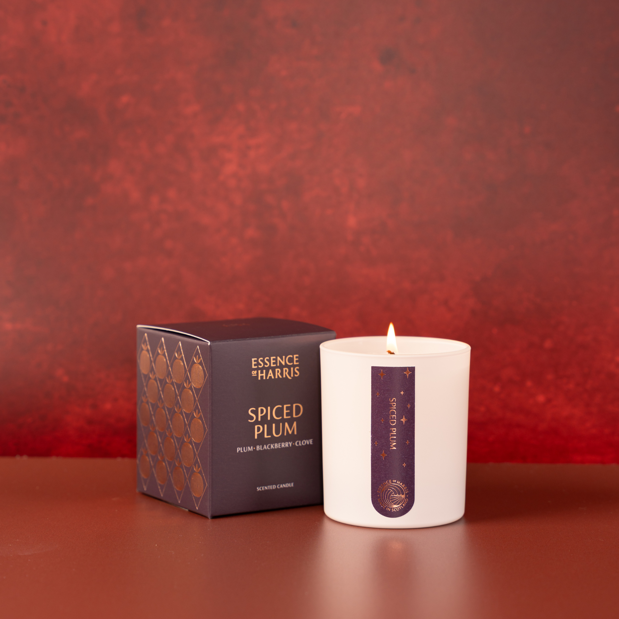 A white glass candle jar with a lit flame sits next to its dark purple box packaging. The packaging features gold foiled text reading 'Essence of Harris' and 'Spiced Plum'.