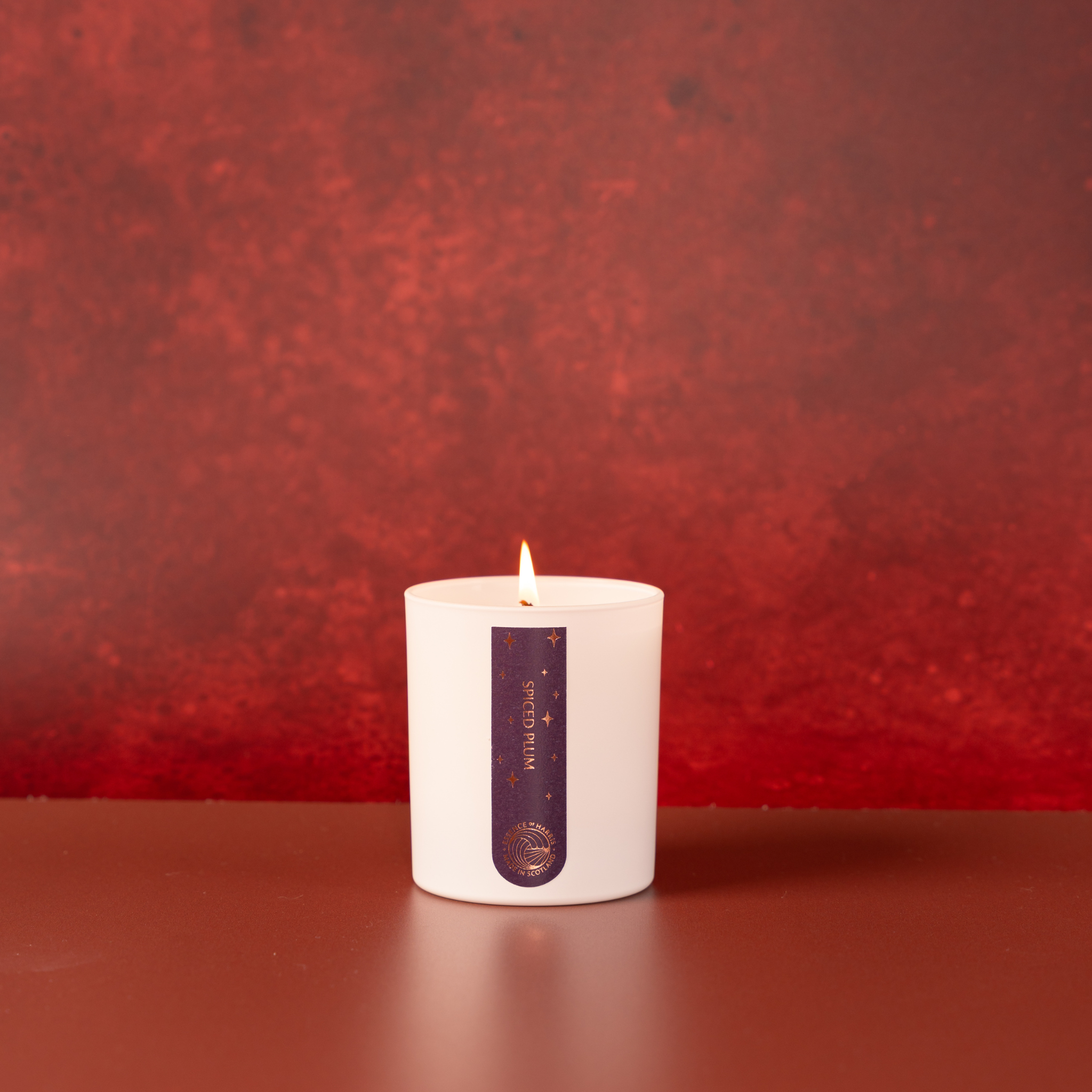 A white jar candle with a lit flame and a vertical dark purple label reading Spiced Plum, set on a brown surface against a textured red background.