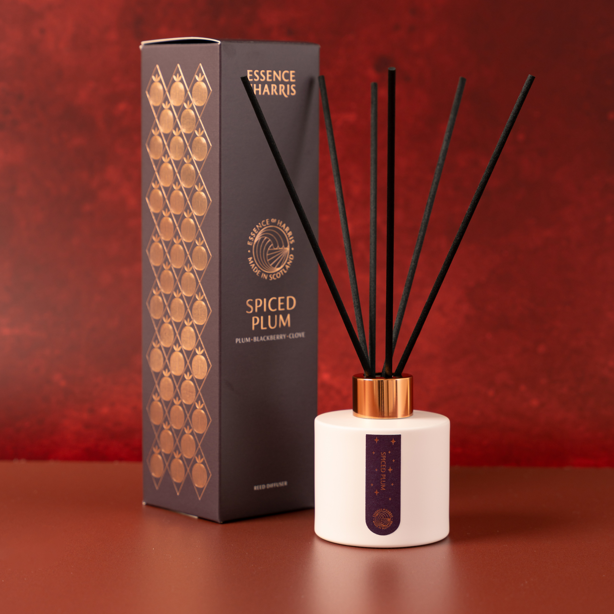 Essence Harris Spiced Plum reed diffuser with a white ceramic bottle, black reeds, a gold cap, and its dark gray packaging box with gold patterns, set against a textured red background.