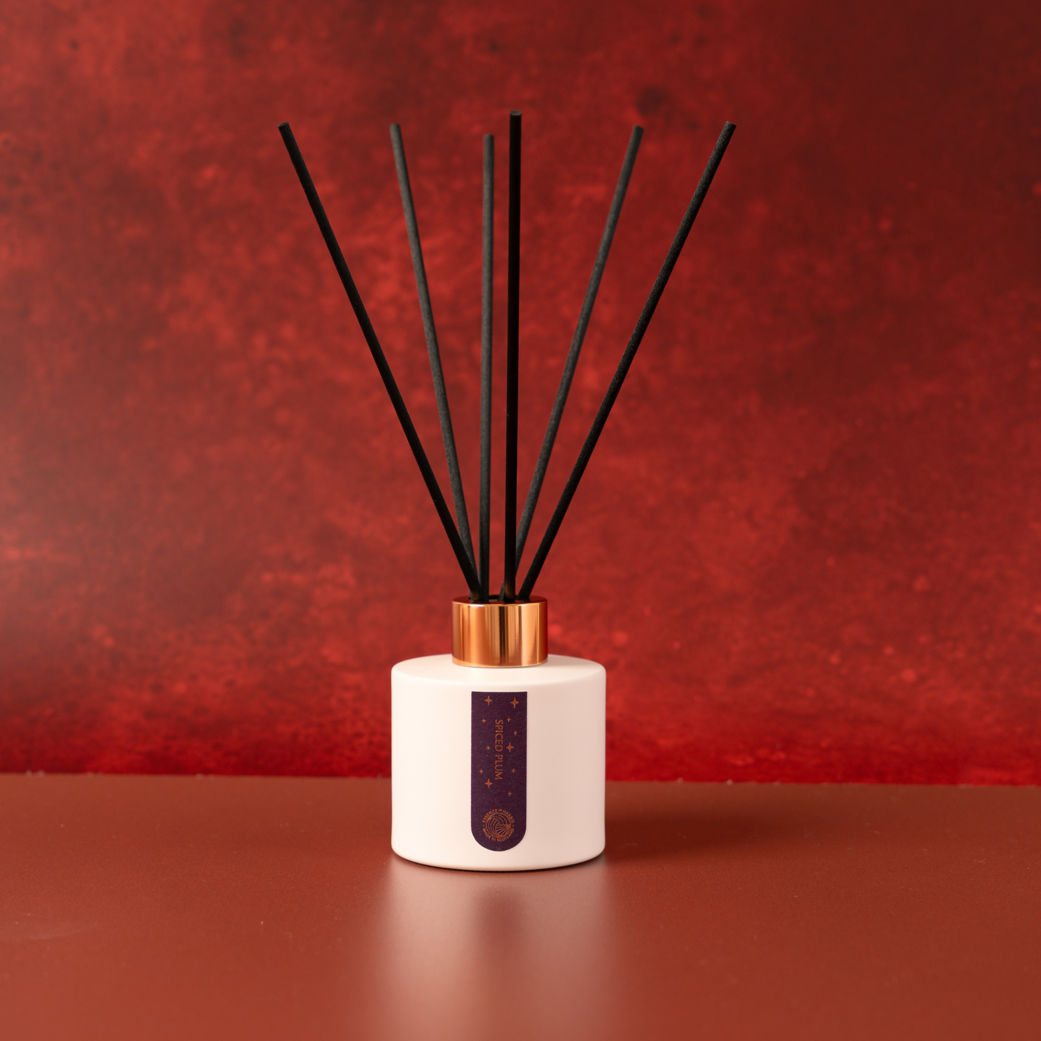 Essence Harris Spiced Plum reed diffuser with a white ceramic bottle, black reeds, a gold cap, set against a textured red background.