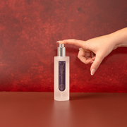 A frosted glass bottle of Essence of Harris Spiced Plum Room Spray with a silver pump dispenser and a dark purple label, set on a matching reddish-brown surface against a textured red background. A hand with manicured nails is reaching toward the pump.