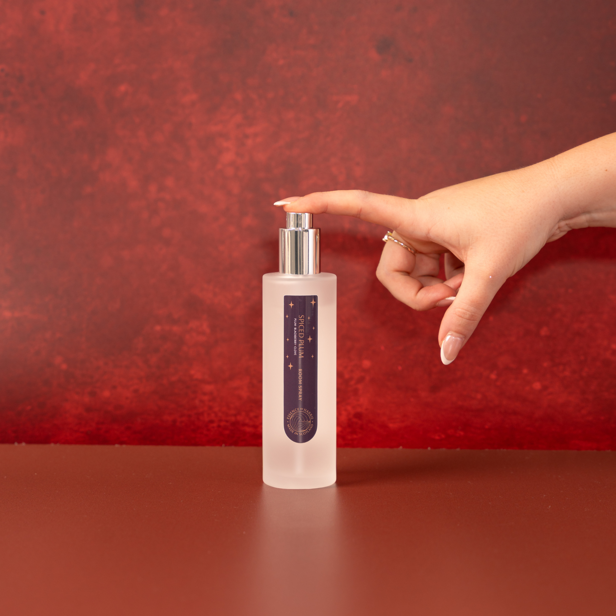 A frosted glass bottle of Essence of Harris Spiced Plum Room Spray with a silver pump dispenser and a dark purple label, set on a matching reddish-brown surface against a textured red background. A hand with manicured nails is reaching toward the pump.